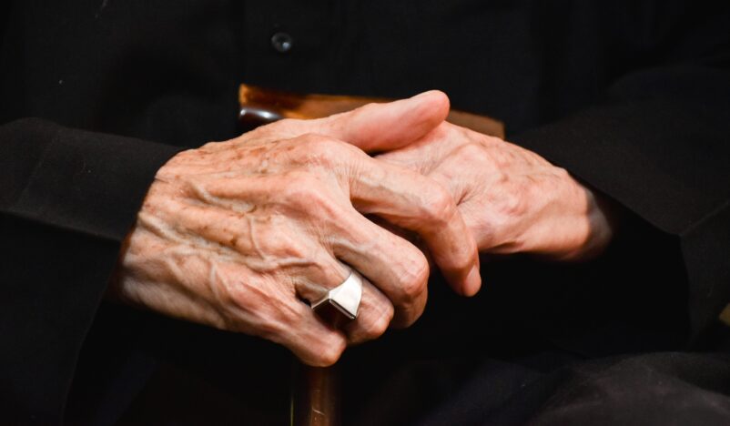 Bild på äldre persons händer. Ring på höger ringfinger. Svart bakgrund.